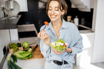 Young, thin, and cheerful woman eating healthy salad in the kitchen at home.