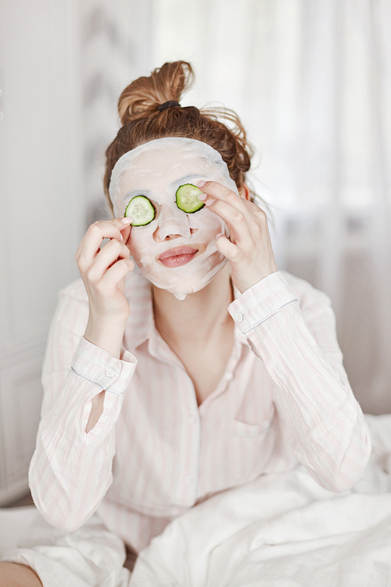 Young woman with face mask treatment putting cucumbers on eyes.