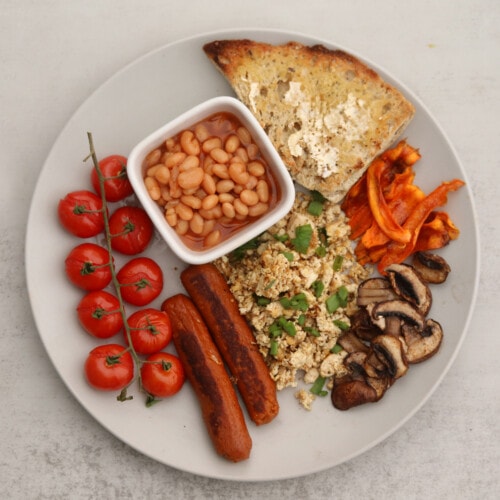 A plate of vegan English breakfast, with scrambled tofu, vegan sausage, baked beans, toast, and roasted tomatoes, mushrooms, and carrots.