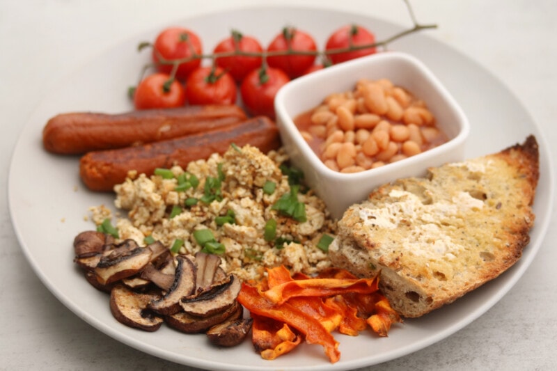 A plate of vegan English breakfast, with scrambled tofu, vegan sausage, baked beans, toast, and roasted tomatoes, mushrooms, and carrots.