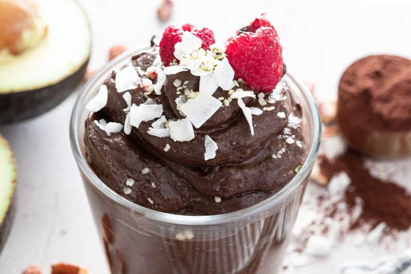 Vegan avocado chocolate pudding in a glass, topped with shaved coconut and raspberries.