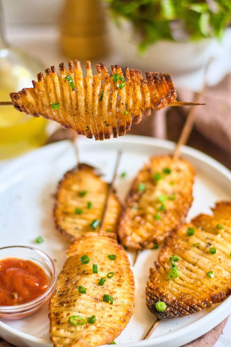 A hand holding a baked accordion potato skewer, with a plate and more potatoes in the background.
