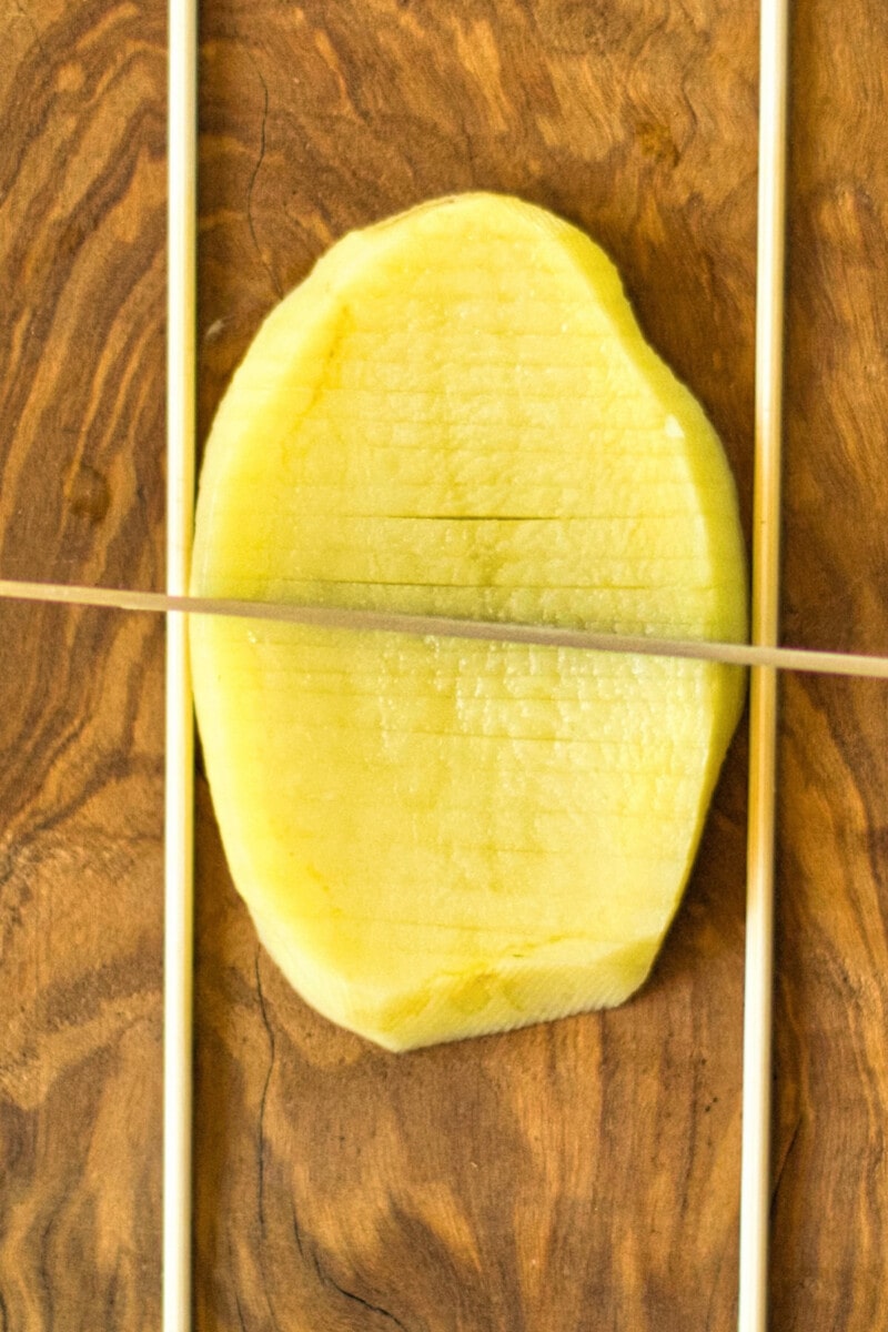 A 1/2" potato slab on a cutting board, cutting shallow, horizontal grooves and using two skewers to prevent cutting all the way through.