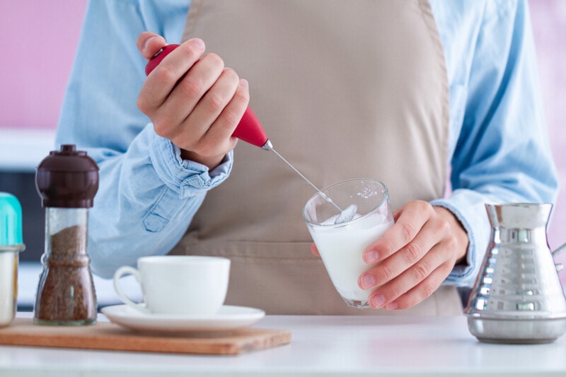 Closeup of a person using a handheld milk frother, frothing a glass of almond milk.