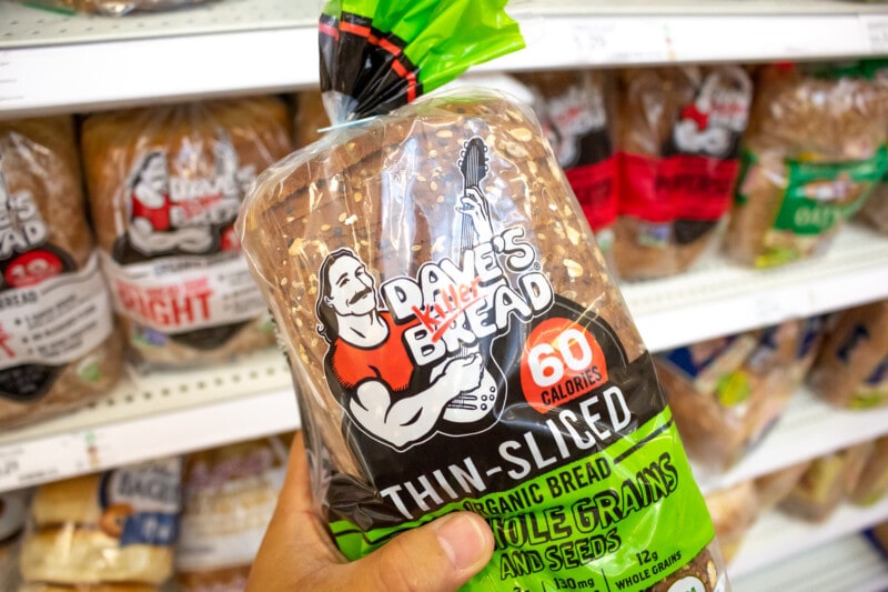 A hand holding a loaf of Dave's Killer Bread in a grocery store aisle.