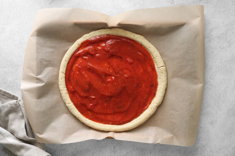 Pizza crust with marinara sauce on parchment paper.