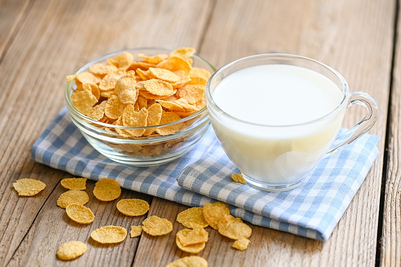 A bowl of cornflake cereal next to a cup of milk on a table.