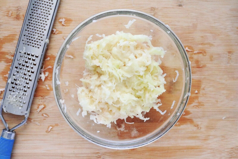 grated potatoes in a glass bowl on a cutting board with a hand grater