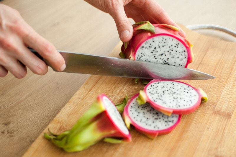 Female hands cutting a dragon fruit or pitaya with pink skin and white pulp with black seeds on wooden cut board on the table.