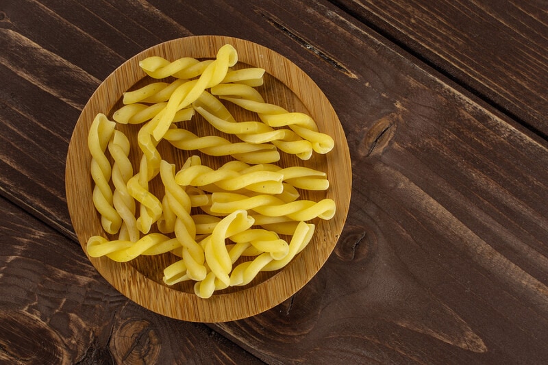 Uncooked gemelli pasta on a bamboo plate on a brown wood table.