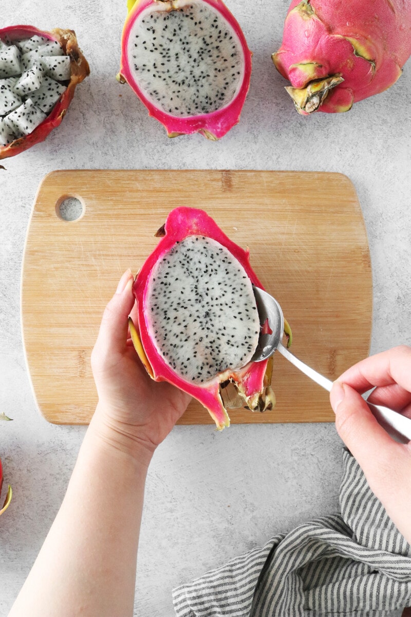 A woman's hands using a spoon to scoop out dragon fruit flesh from the skin.