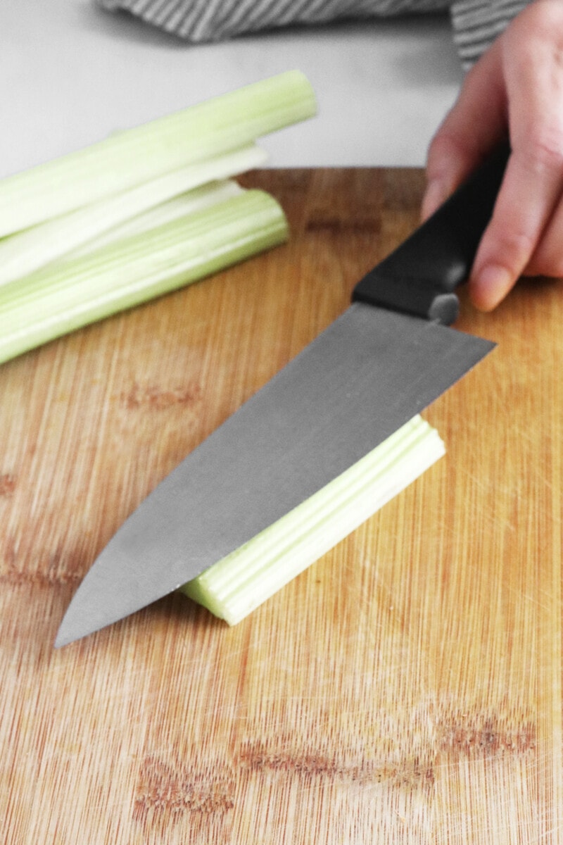 Placing flat side of a large chef's knife on top of celery on a cutting board.