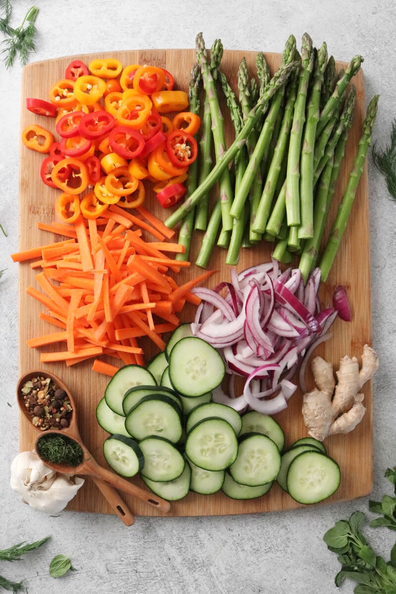 Chopped vegetables on a wooden cutting board, including sliced sweet peppers, red onions, carrots, cucumbers, ginger, and asparagus.