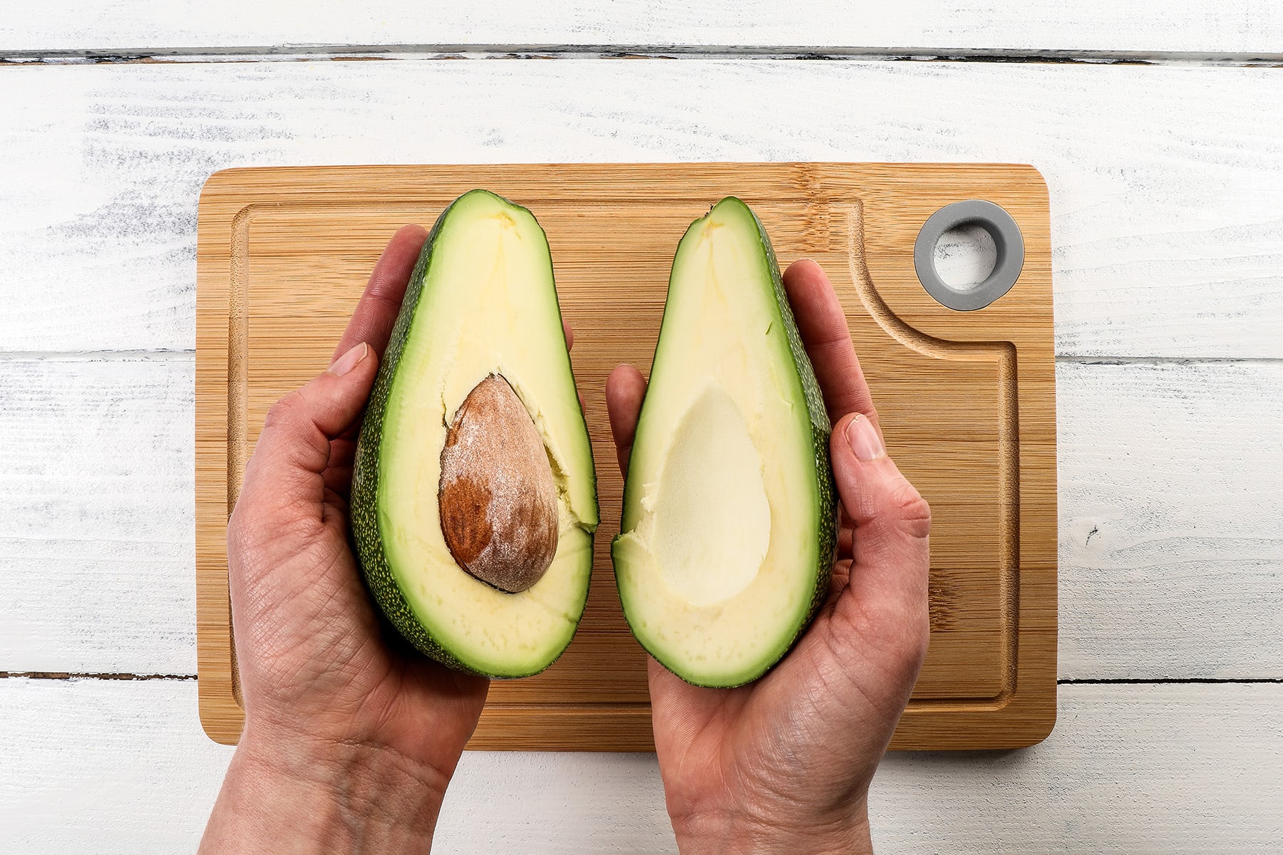 A woman's hands holding two avocado halves over a bamboo cutting board.