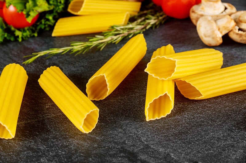 Dry manicotti pasta with mushrooms and tomato on black background.