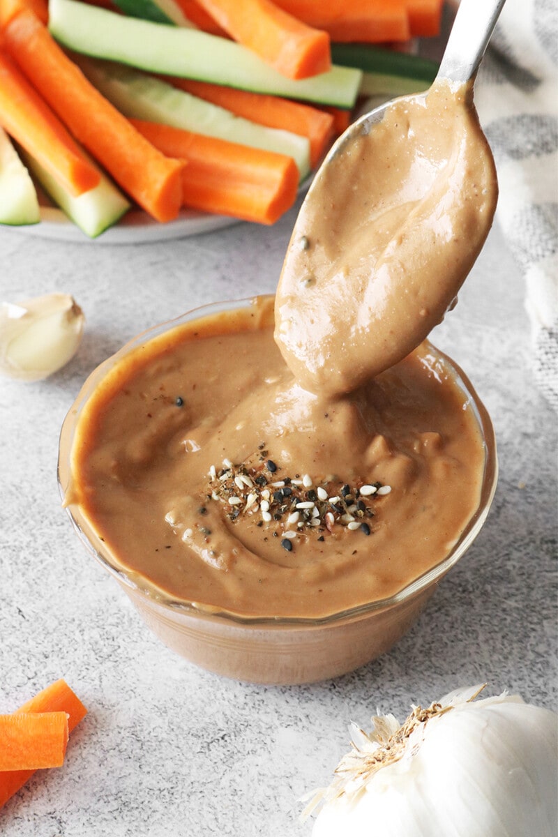 A spoon dipped into a bowl of vegan peanut sauce, topped with sesame seeds.