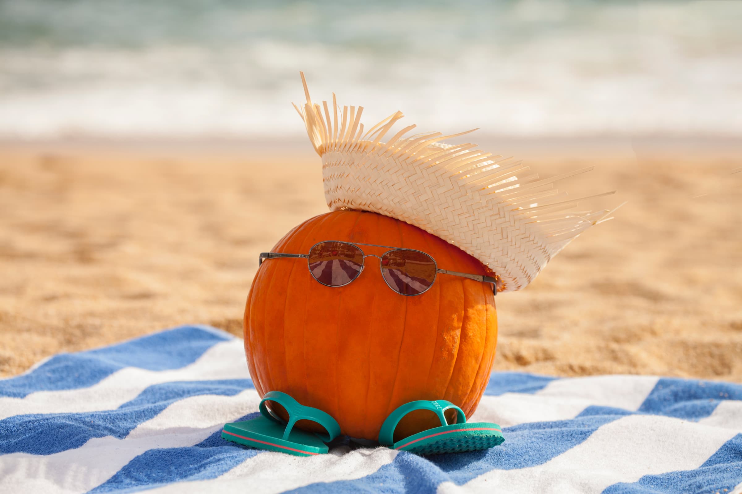 A pumpkin on a towel on a beach wearing sunglasses and a straw hat