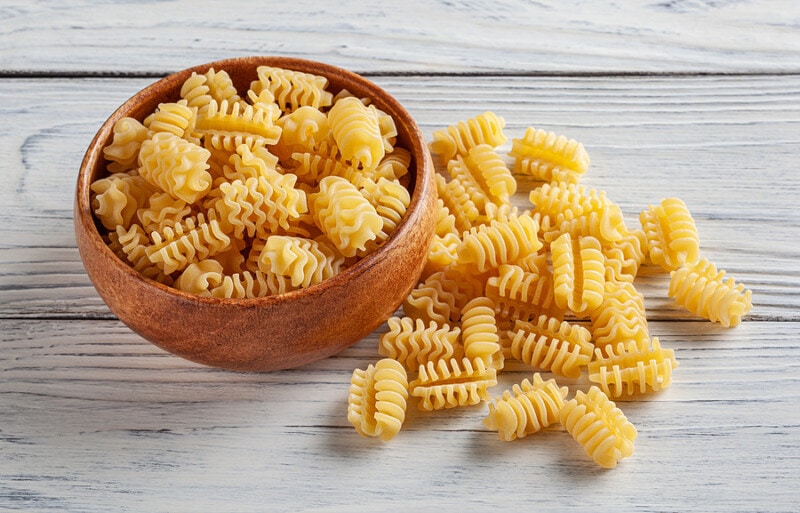 Uncooked radiatori pasta in wooden bowl on white wooden background.