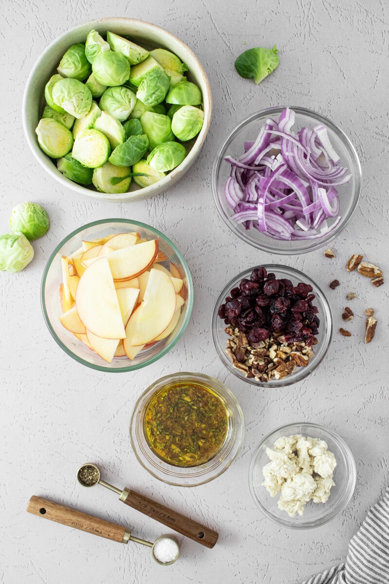 Ingredients for roasted Brussels sprouts salad: fresh brussel sprouts, red onion, thinly sliced apples, dried cranberries, vegan feta, and dressing.