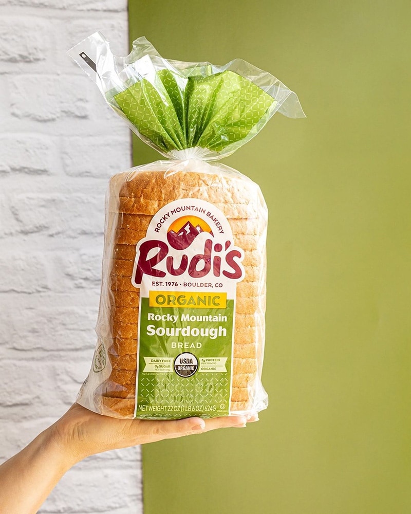 A hand holding a loaf of Rudi's Organic Rocky Mountain Sourdough bread.