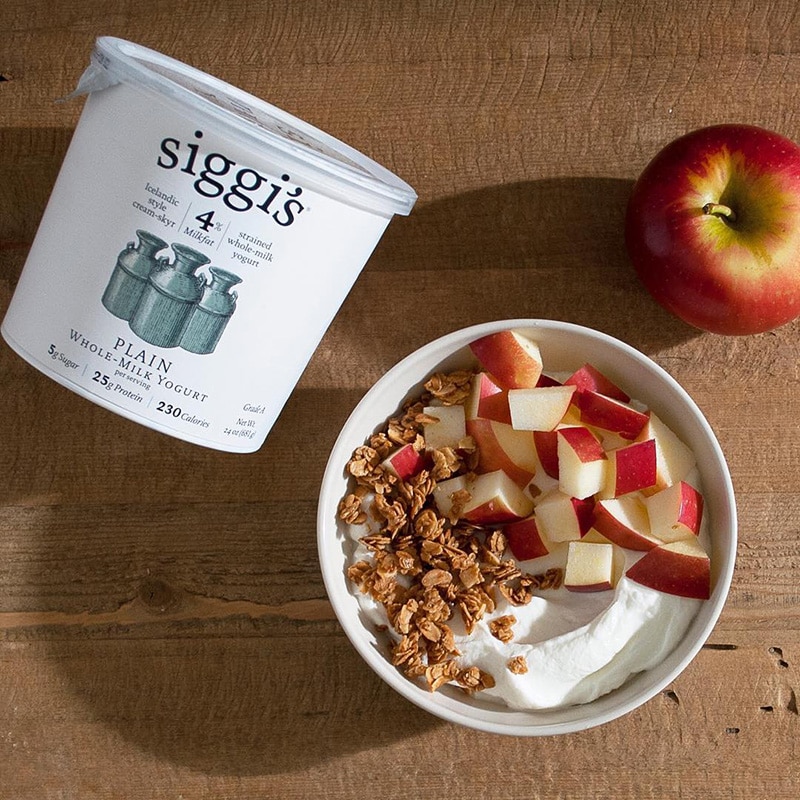 A large container of Siggi's skyr yogurt next to a bowl of yogurt, granola, and cubed apples.