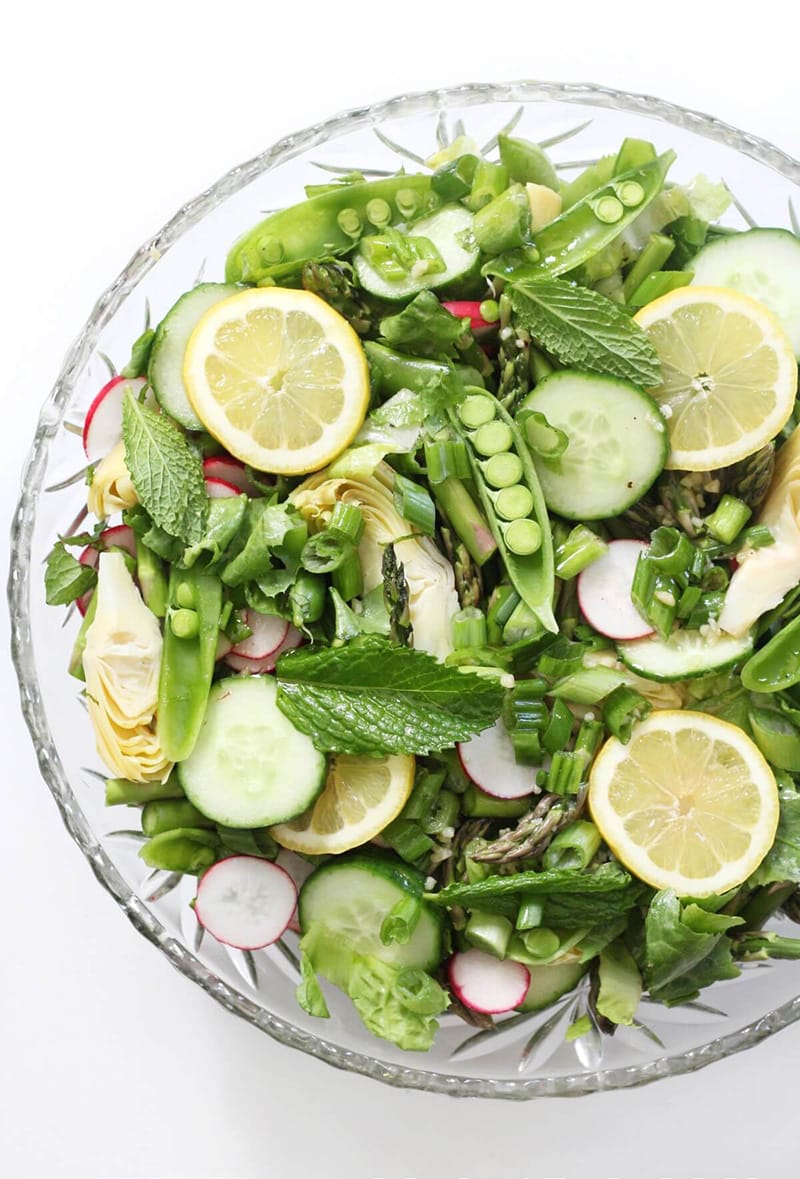 Fresh greens topped with snap peas, sliced cucumbers, radishes, lemon, and mint in a bowl.
