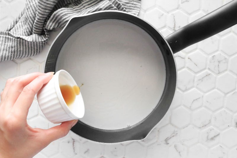 Woman's hand pouring maple syrup into a pot of coconut milk on a white counter