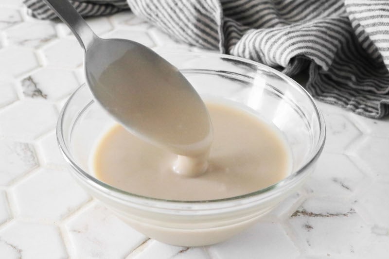 Vegan condensed milk dripping off of a spoon into a small glass bowl on a white counter