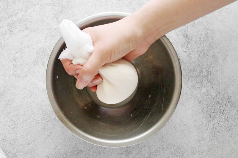 A hand squeezing the yogurt mixture in the cheese cloth above a bowl to strain.