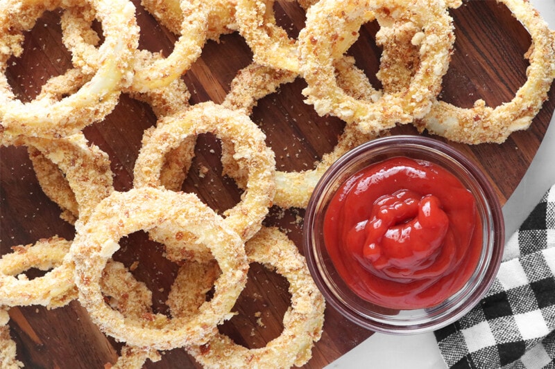 Vegan crispy baked onion rings on a serving board served with a side of ketchup.