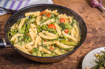 Cooking primavera pasta in a pan - vegan american and italian primavera pasta dish with broccoli, beans, asparagus, peas and tomatoes in a pan on a wooden table, top view.
