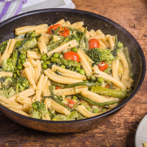 Cooking primavera pasta in a pan - vegan american and italian primavera pasta dish with broccoli, beans, asparagus, peas and tomatoes in a pan on a wooden table, top view.