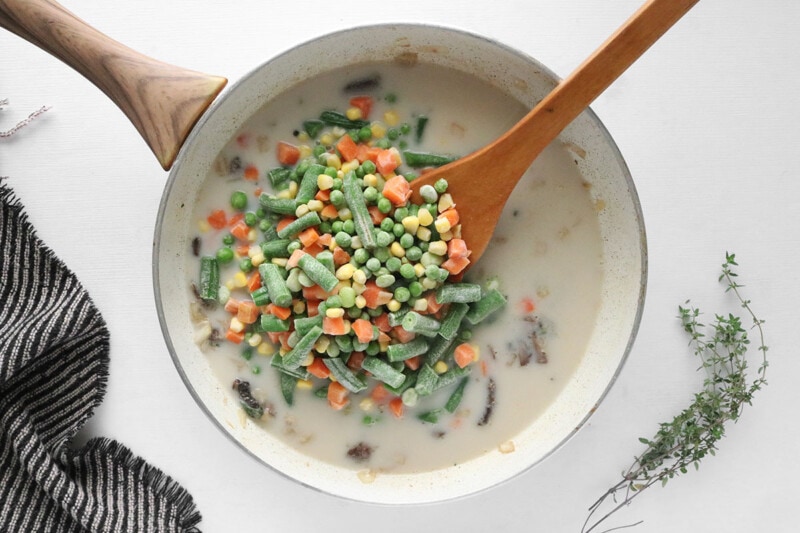Frozen vegetables being added to a mushroom and onion mixture in a skillet with a wooden spoon