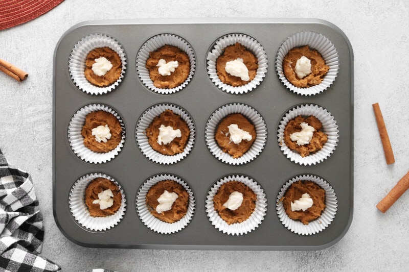 Muffin pan lined with muffin cups and filled with batter and a dollop of vegan cream cheese mixture.