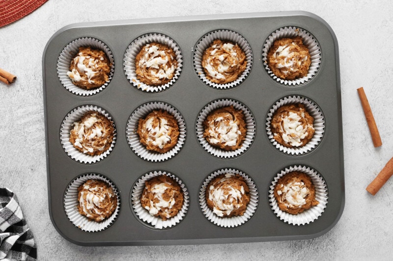 Muffin pan lined with muffin cups and filled with batter and vegan cream cheese swirled on top.
