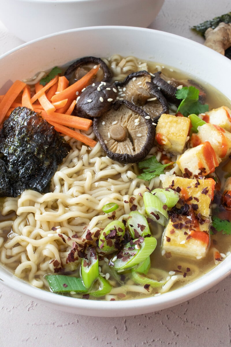 Large bowl of vegan ramen soup, topped with shiitake mushrooms, carrots, tofu, scallions, and nori.