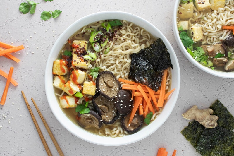 Large bowl of vegan ramen soup, topped with shiitake mushrooms, carrots, tofu, scallions, and nori.
