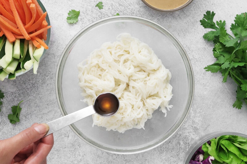 Tossing cooked rice noodles with sesame oil in a large bowl.