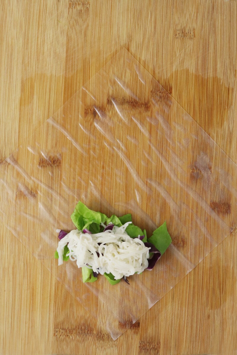 Rice paper positioned on a bamboo cutting board in the shape of a diamond, with rice noodles, cabbage, and lettuce placed on top, one inch from the bottom corner of the paper.