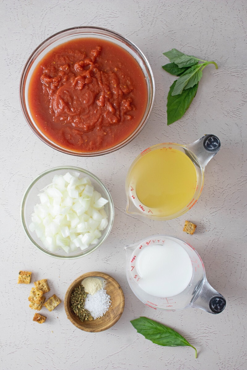 Ingredients for vegan tomato soup: Crushed tomatoes, vegetable broth, onion, plant-based milk, fresh basil, and spices.