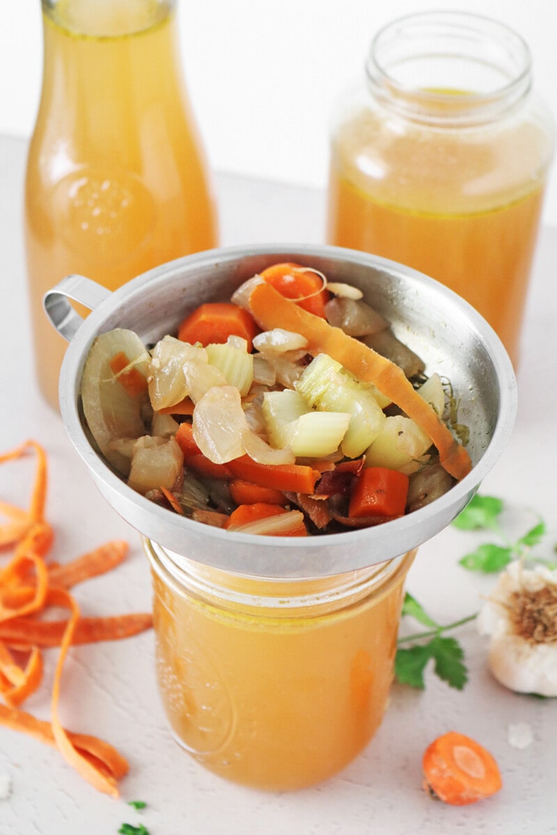 Vegan vegetable broth with carrots, onions, leeks, and fresh herbs in glass jars.