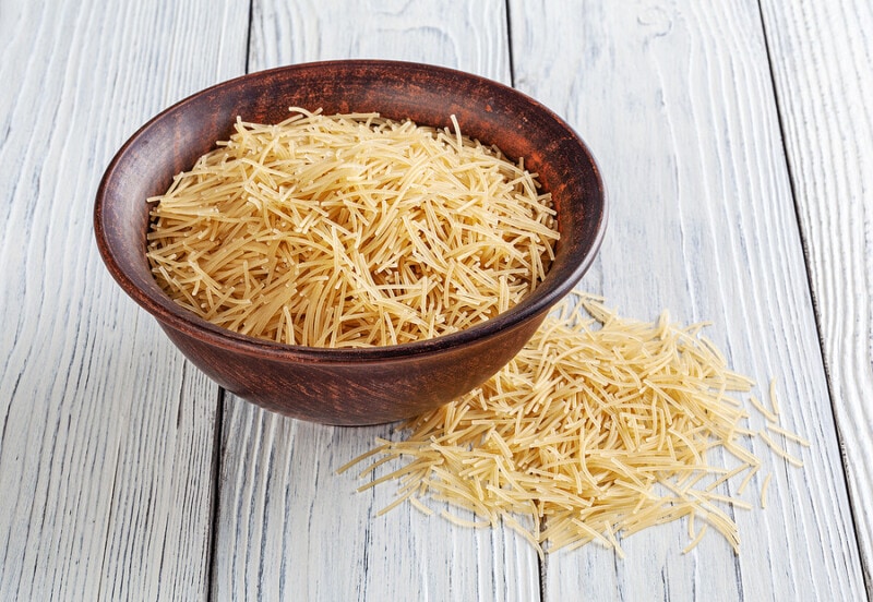 Uncooked vermicelli pasta in ceramic bowl on white wooden background.