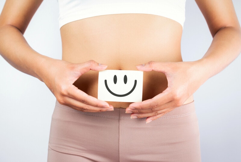 Woman in white top with exposed midriff and mauve leggings holding a paper happy face with her hands in front of her stomach.