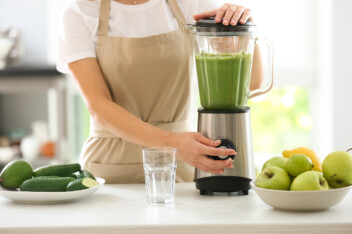 woman in tan apron using a blender to make a green smoothie